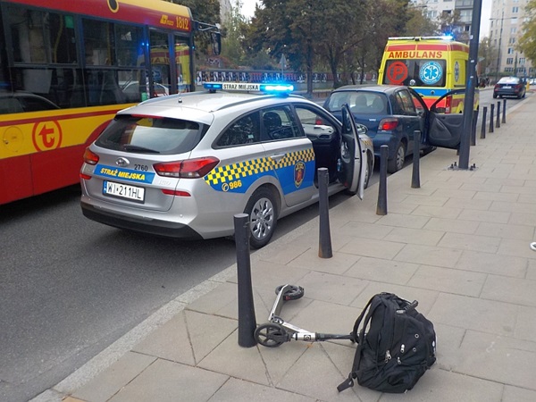 Samochód policyjny na miejscu wypadku ze skuterem i plecakiem; w tle karetka pogotowia i autobus