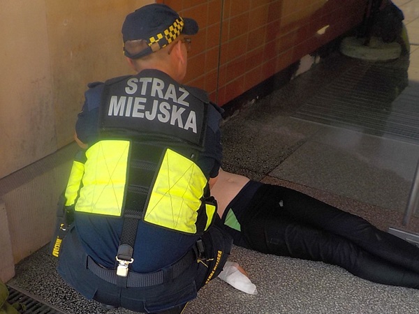 Funkcjonariusz Straży Miejskiej pomaga osobie leżącej na ziemi.