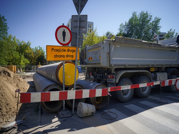 Plac budowy z dużymi rurami, wywrotką i znakiem na przejściu dla pieszych z napisem „Przejście druga strona drogi”.