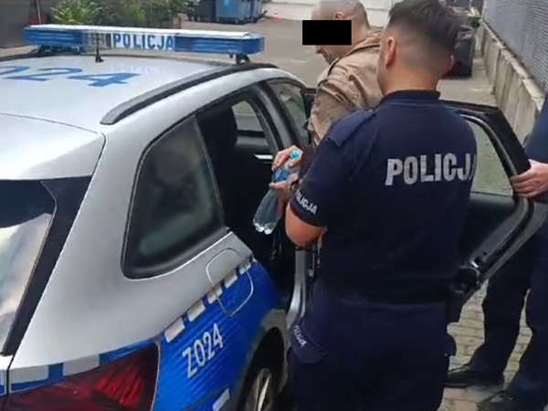 Polscy policjanci zatrzymują podejrzanego w radiowozie. Na radiowozie i mundurze policjanta widoczny jest napis „POLICJA”.