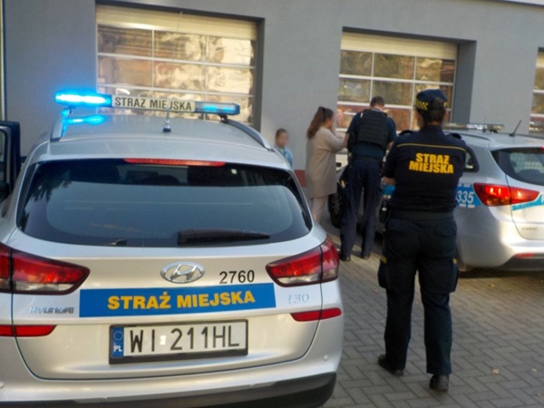 Dwóch polskich funkcjonariuszy Straży Miejskiej rozmawia z rodziną przy radiowozie. Na pojeździe widoczny jest napis „Straż Miejska”.