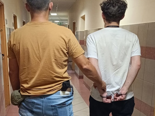 Dwóch mężczyzn w kajdankach, jeden w koszuli w kolorze musztardowym, a drugi w białej koszuli, idących korytarzem. Aresztowanie.