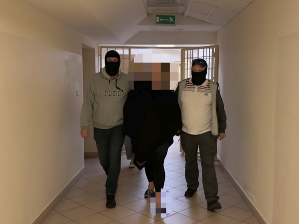 Trzy osoby w maskach eskortowane korytarzem. Jedna osoba jest w środku, otoczona przez dwie inne.