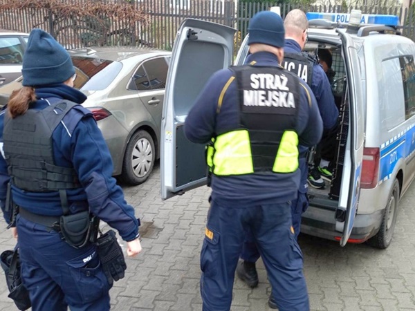Policjanci miejscy zatrzymują podejrzanego w pobliżu samochodu. Na kamizelce funkcjonariusza widać napis „Straż Miejska”.