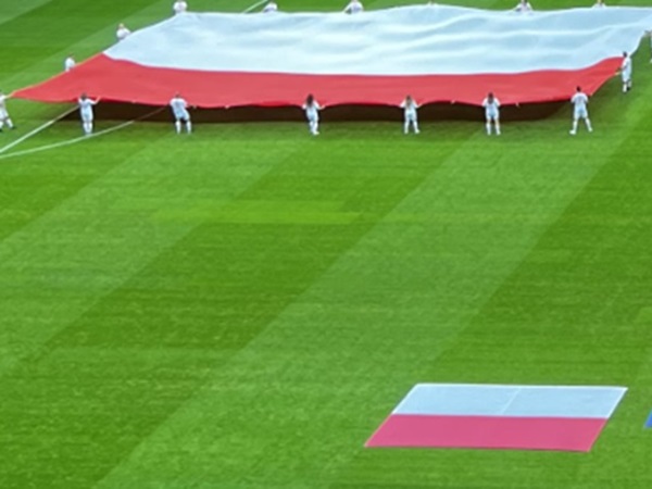 Ludzie ubrani na biało rozwijają dużą polską flagę na boisku piłkarskim.