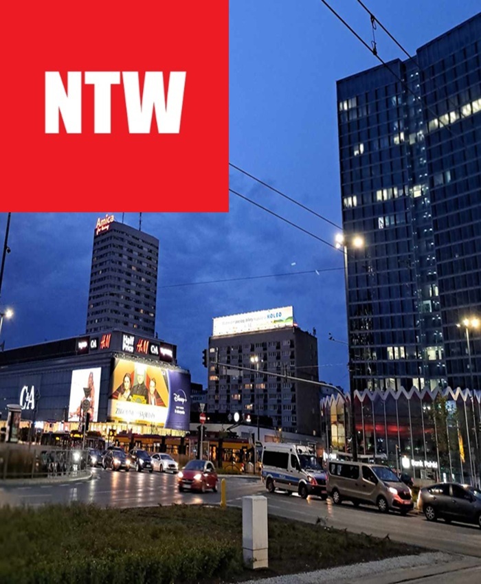 Nocny widok skrzyżowania ulic miasta, z nowoczesnymi budynkami i billboardami, w tym logo NTW.
