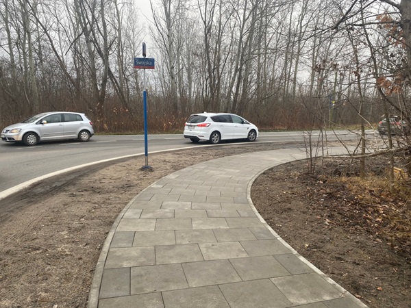 Zakrzywiony chodnik przy drodze z samochodami i znakiem przystanku autobusowego z napisem „Swojska”.
