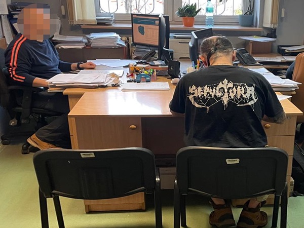 Dwóch mężczyzn pracuje przy biurkach, jeden z nich ma na sobie czarną koszulkę z nadrukiem zespołu Servus Dei.