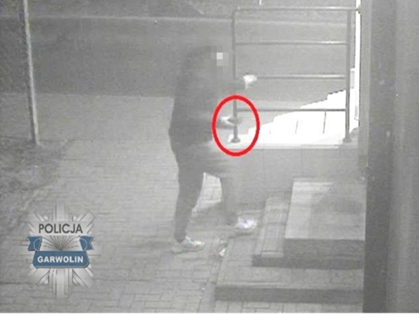 Obraz z monitoringu przedstawiający podejrzanego używającego łomu w pobliżu budynku.