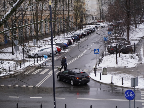 Piesi przechodzący przez zaśnieżoną ulicę w mieście, samochody zaparkowane wzdłuż krawężnika.