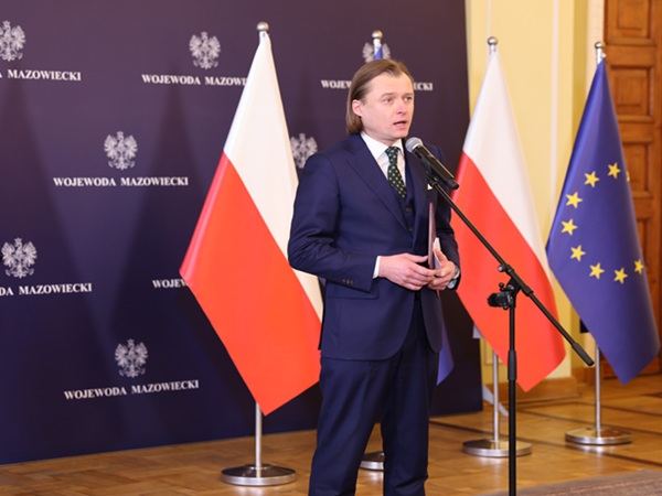 Polski urzędnik przemawiający na konferencji prasowej. Widoczne flagi Polski i UE.