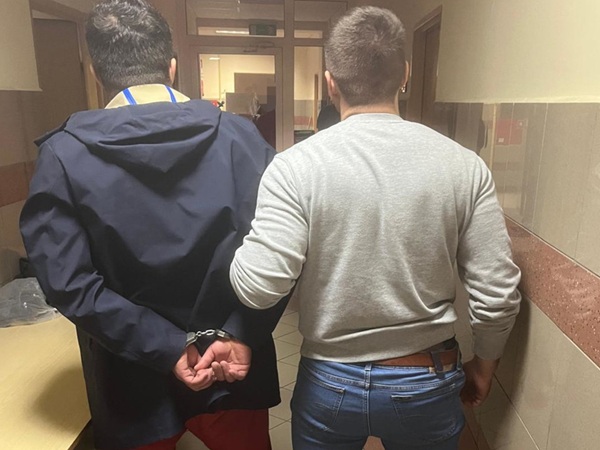 Dwóch mężczyzn w kajdankach, widok z tyłu, idących korytarzem.