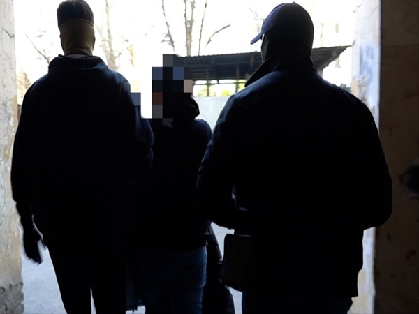 Trzy osoby w ciemnych ubraniach wchodzą przez drzwi, twarz jednej osoby jest niewyraźna.