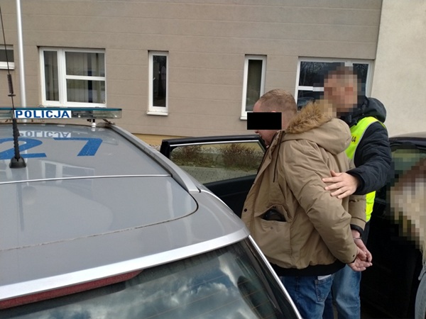 Policja aresztuje podejrzanego w kajdankach w pobliżu radiowozu.