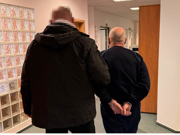 Dwóch mężczyzn w kajdankach, jeden w ciemnej kurtce, idących korytarzem.