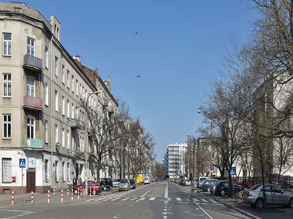 Scena uliczna w Warszawie, ukazująca budynki mieszkalne i zaparkowane samochody.