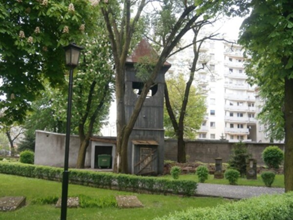 Stara drewniana dzwonnica na spokojnym cmentarzu, otoczona drzewami i nagrobkami.