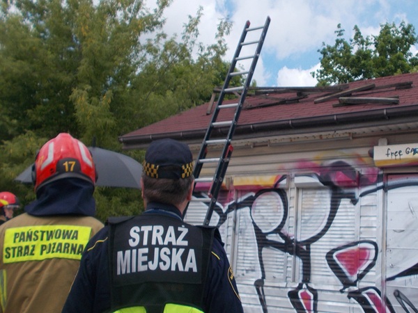 Strażacy oceniają uszkodzenia dachu, stanowiące poważne zagrożenie dla zdrowia i życia. „Straż Miejska” widoczna na mundurze.