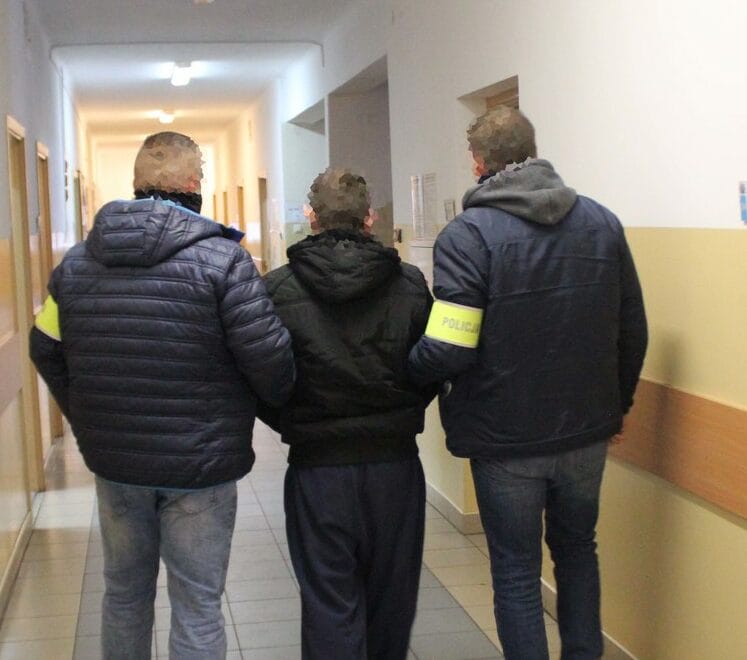 Dwóch policjantów eskortuje podejrzanego korytarzem szpitalnym.