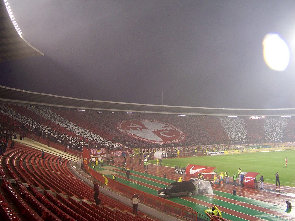 Stadion nocą wypełniony kibicami machającymi czerwonymi i białymi flagami; widoczny baner „Belgrade Boys”.