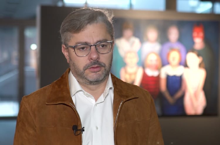 Mężczyzna w brązowej kurtce rozmawia o sztuce na otwarciu galerii.