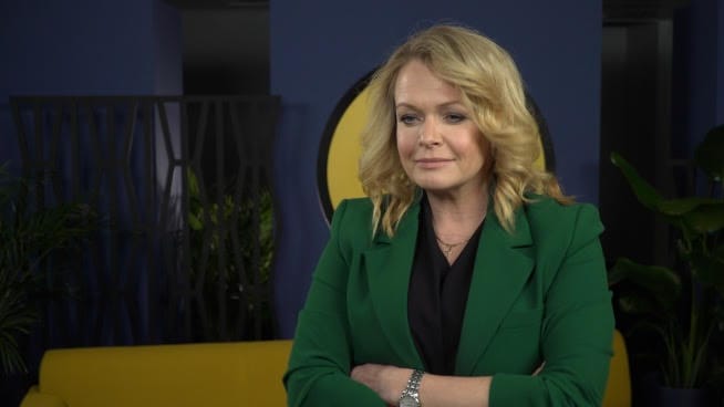 Kobieta w zielonym żakiecie, ze skrzyżowanymi ramionami i zamyślonym wyrazem twarzy.