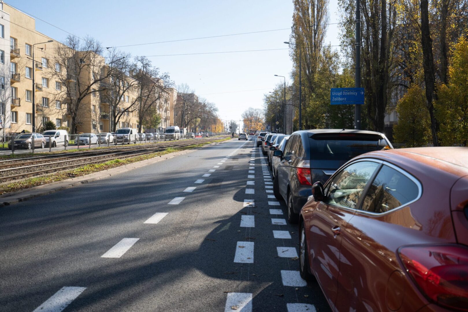 Samochody zaparkowane wzdłuż ulicy miejskiej z torami tramwajowymi, widoczny znak „Urząd Dzielnicy Żoliborz”.