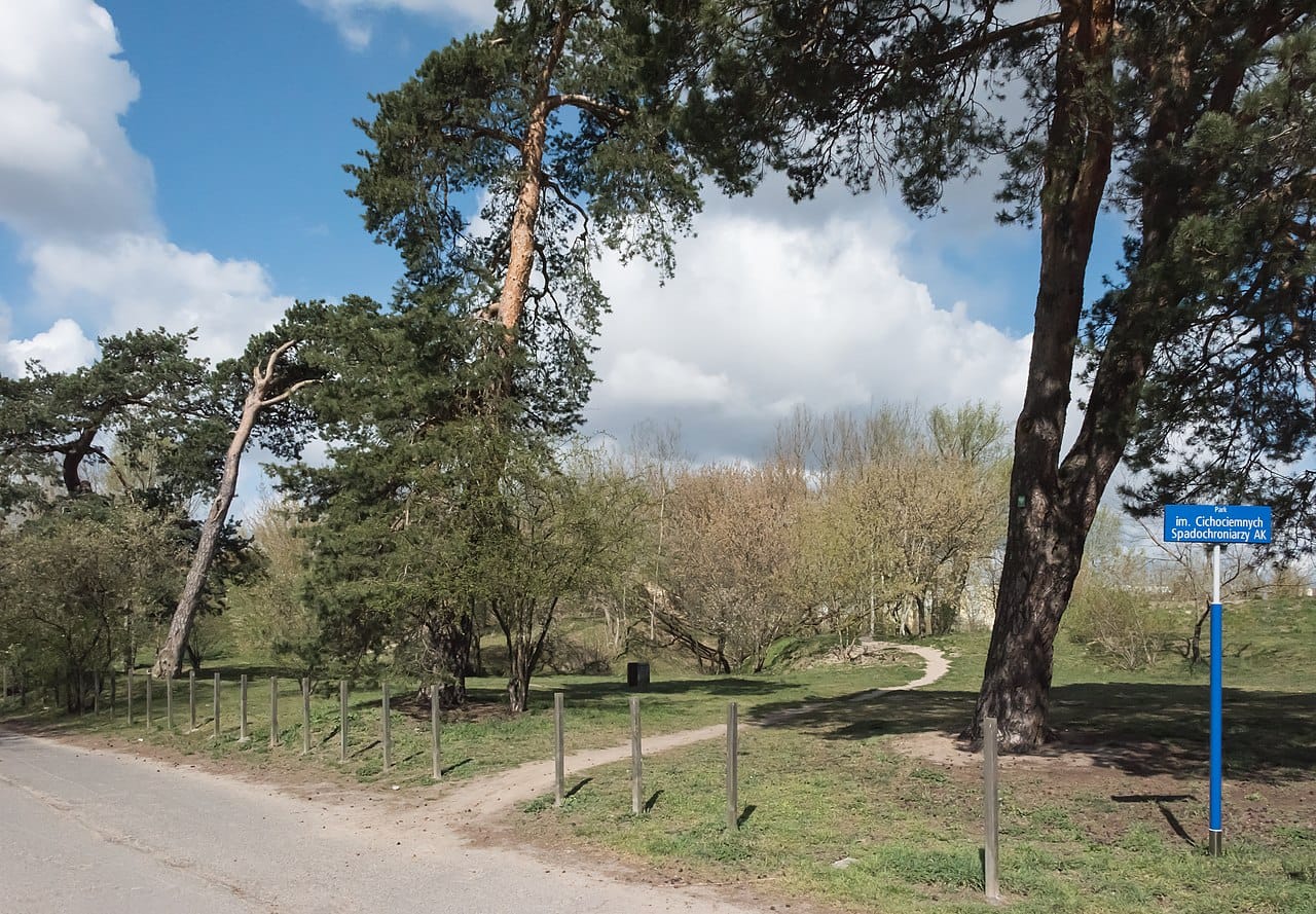 Wejście do parku ze ścieżką, drzewami i znakiem: Park im. Cichociemnych Spadochroniarzy AK