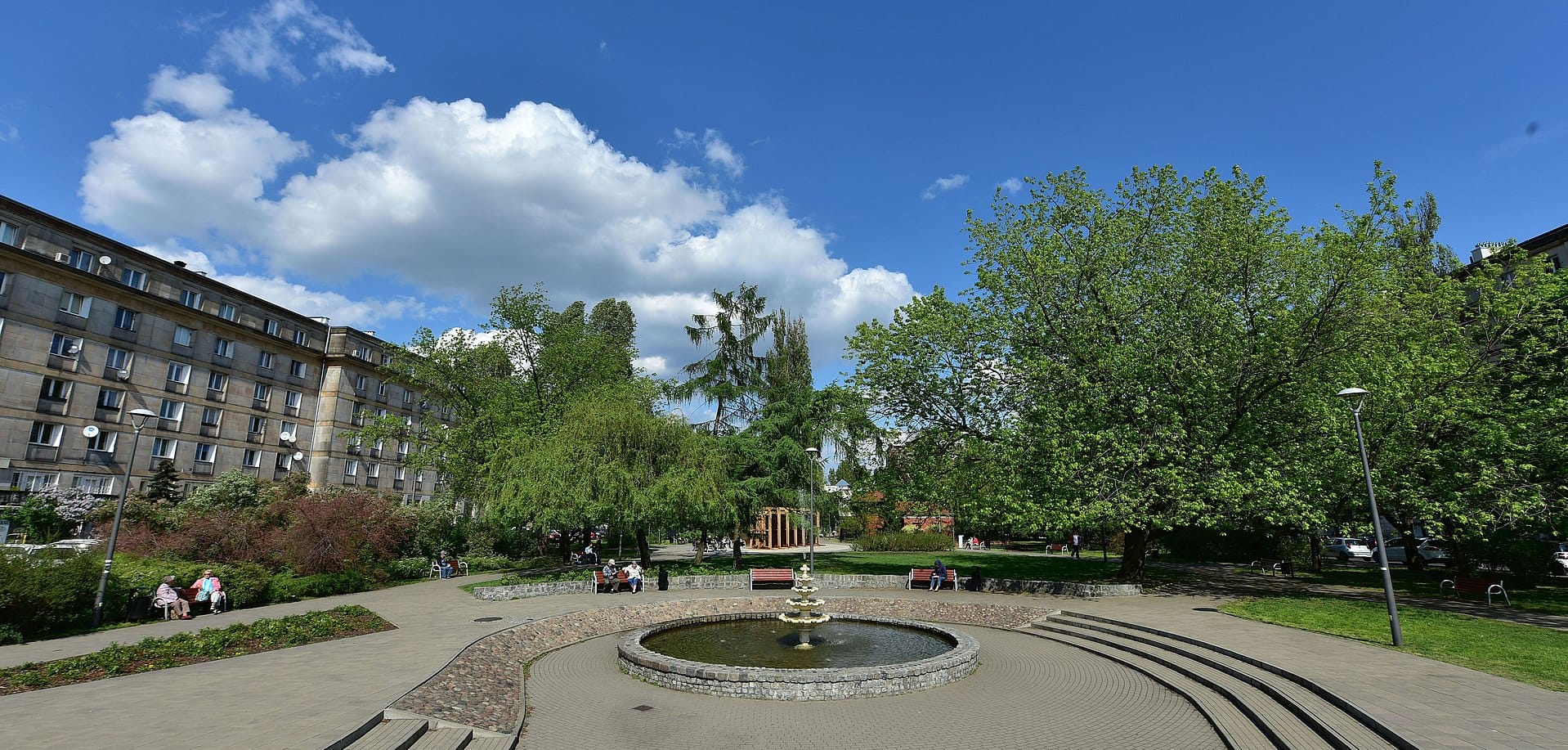 Park miejski z fontanną, ławkami i budynkami mieszkalnymi.