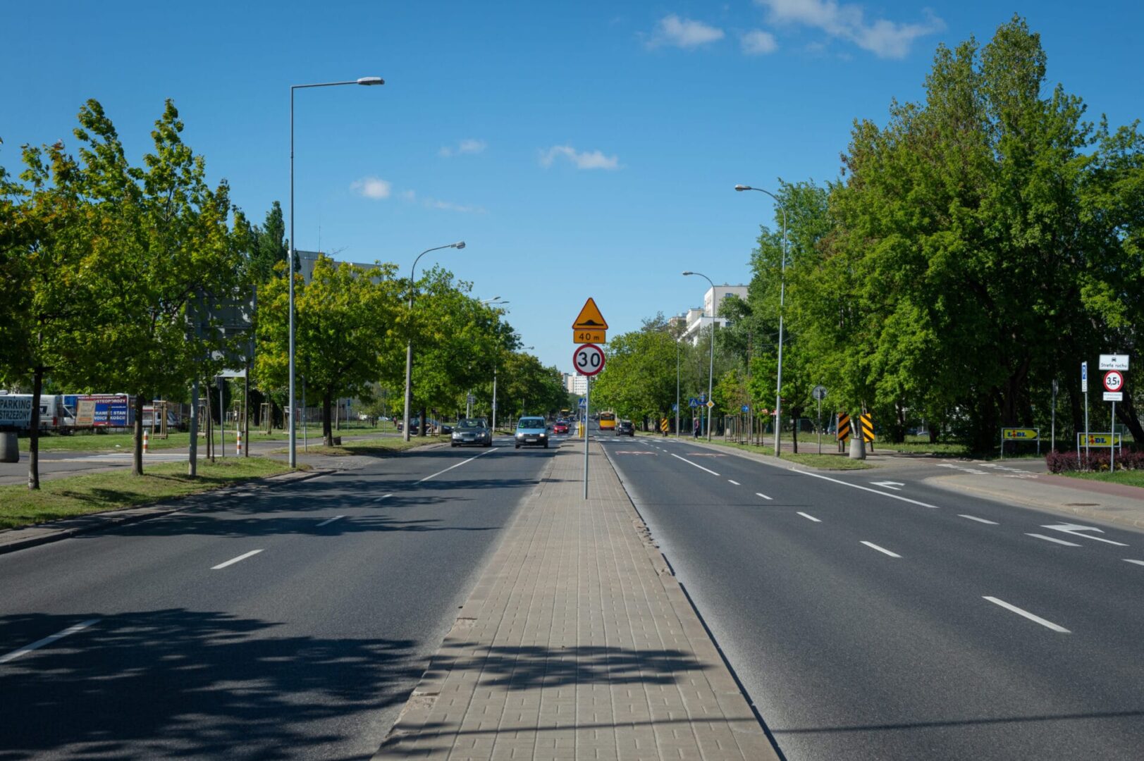 Pusta ulica miejska ze znakiem ograniczenia prędkości do 30 km/h i drzewami rosnącymi wzdłuż drogi.