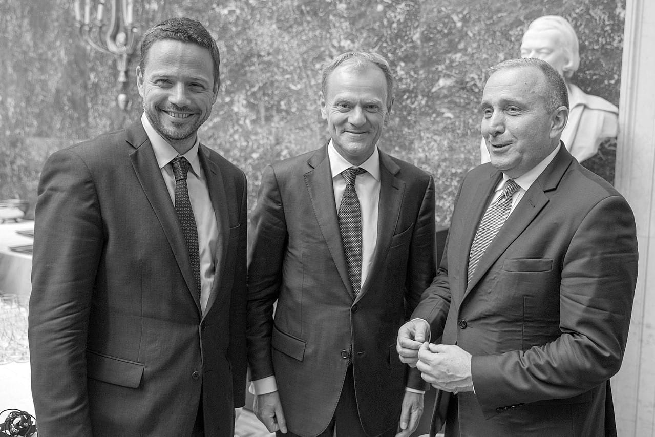 Trzech mężczyzn w garniturach, prawdopodobnie na spotkaniu biznesowym lub konferencji.