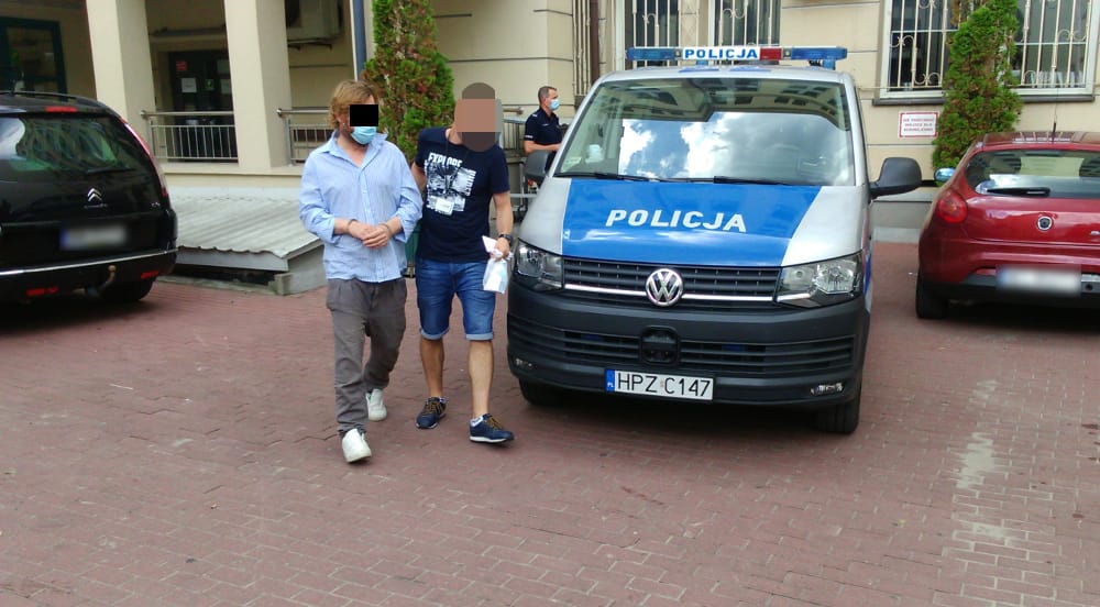 Dwóch mężczyzn w kajdankach eskortowanych przez polską policję. Na radiowozie widać napis „Policja”.