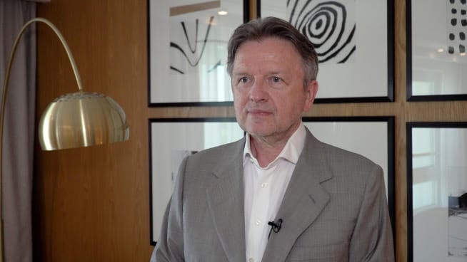 Portret mężczyzny w szarym garniturze na tle ściany z oprawionym obrazem.