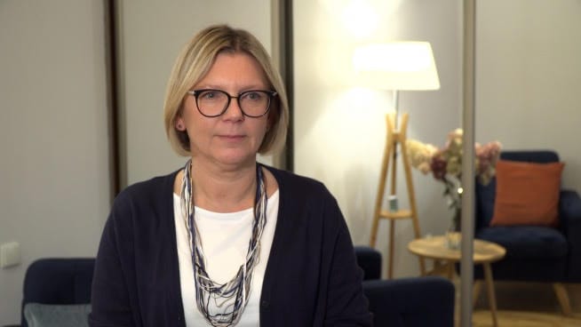 Portret kobiety w okularach i z naszyjnikiem, w otoczeniu profesjonalnym.