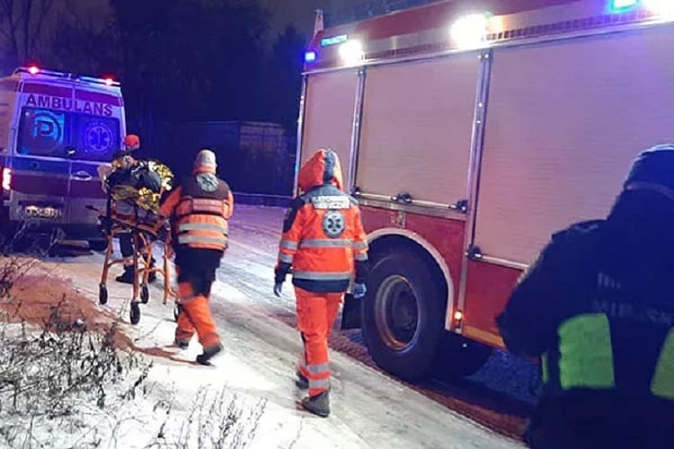 Ratownicy medyczni udzielają pomocy rannej osobie na zaśnieżonej drodze w pobliżu karetki pogotowia i wozu strażackiego.