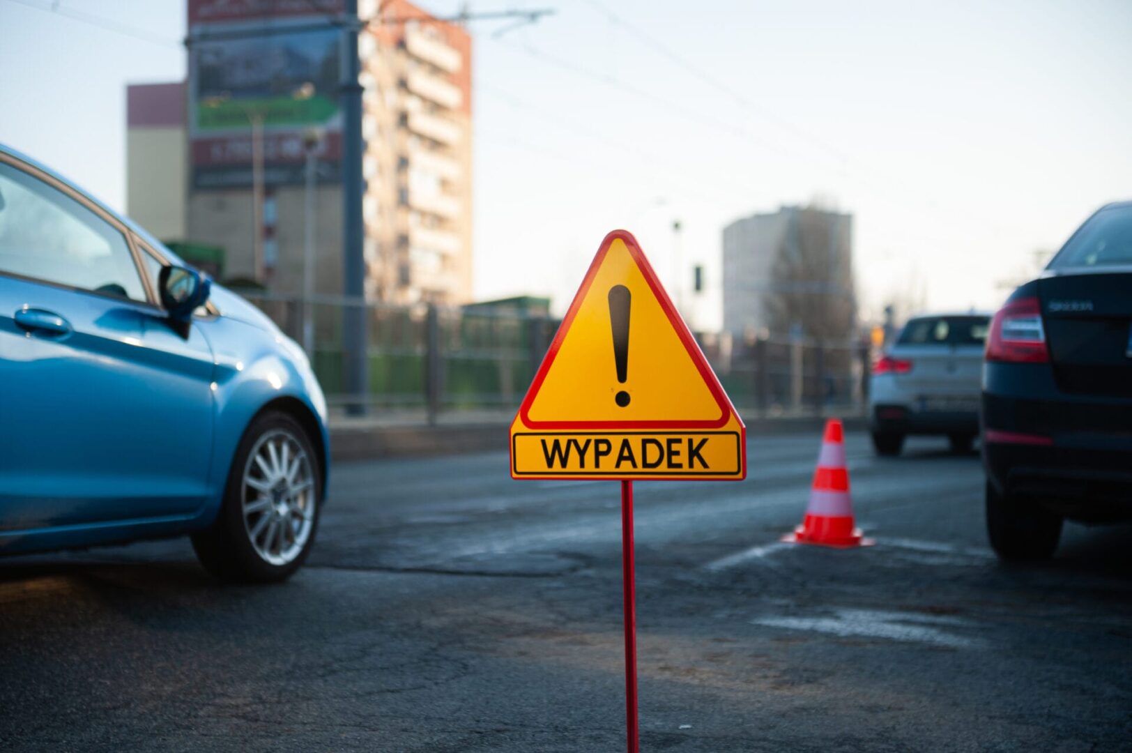 Miejsce wypadku ze znakiem ostrzegawczym "WYPADEK" i uszkodzonymi pojazdami.
