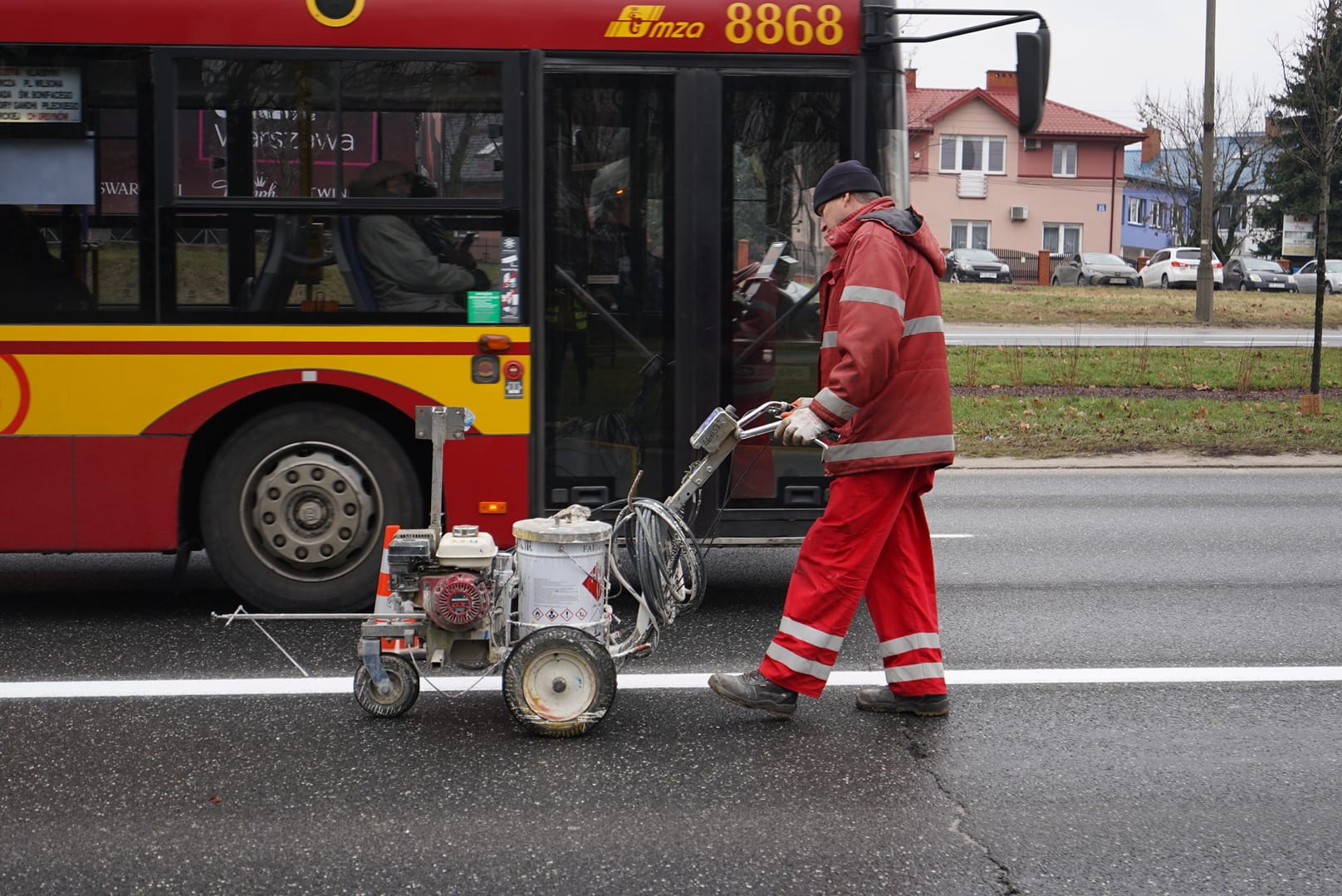 Pracownik malujący czerwone linie drogowe za pomocą maszyny do malowania pasów obok autobusu.