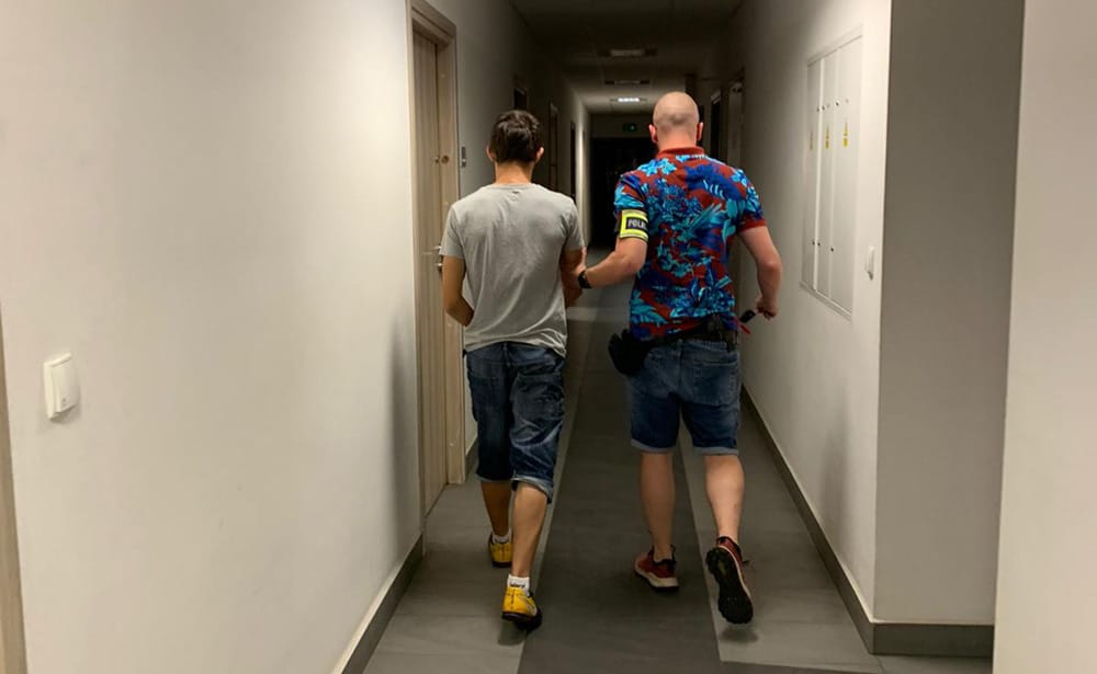 Dwóch mężczyzn idących korytarzem, jeden z nich wygląda na policjanta eskortującego drugiego.