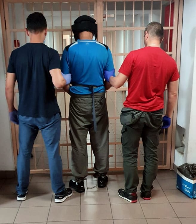 Więzień w kajdanach eskortowany przez strażników przez bramę więzienia