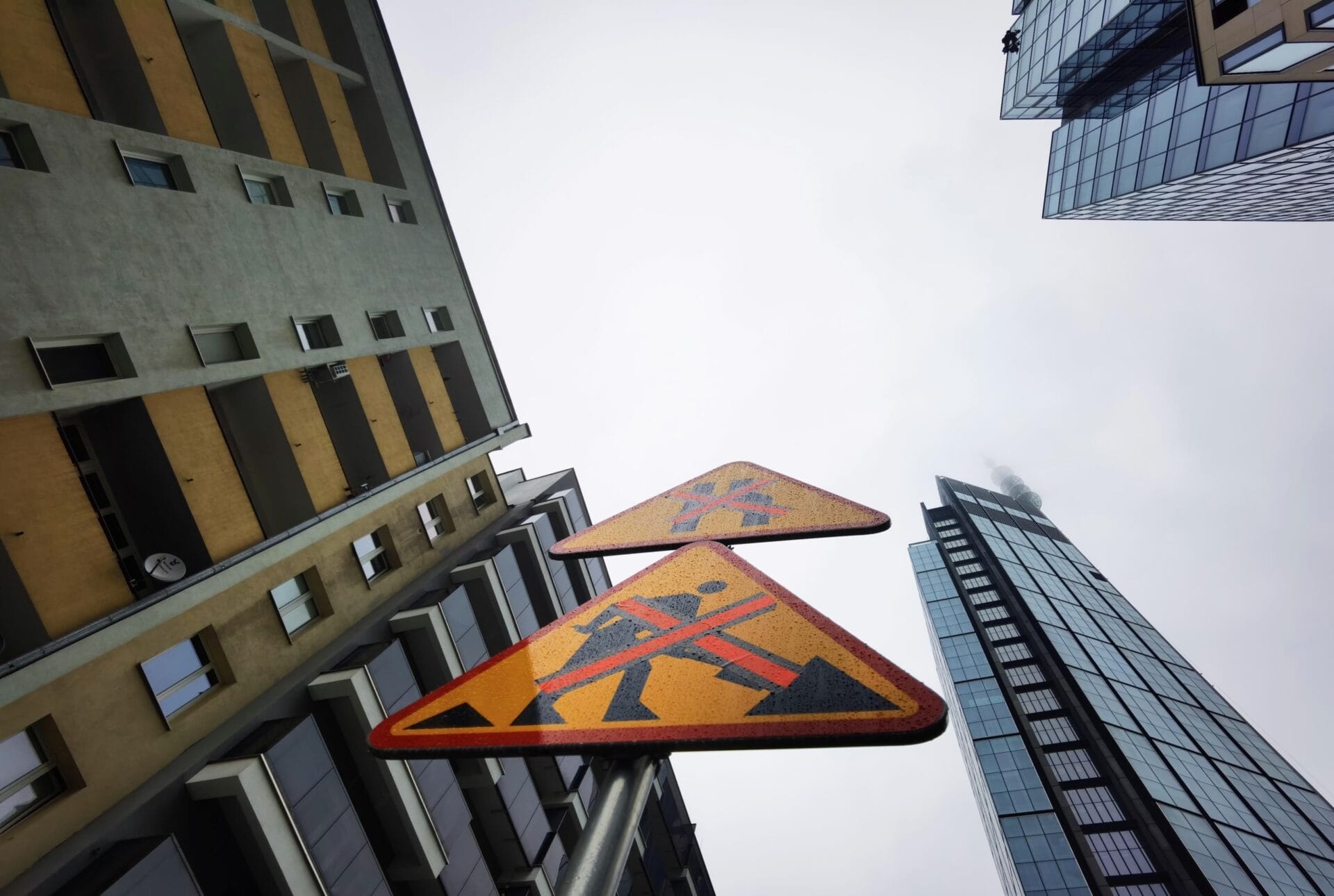 Dwa trójkątne znaki drogowe pokazujące zamknięte przejście dla pieszych, widziane z dołu między wysokimi budynkami