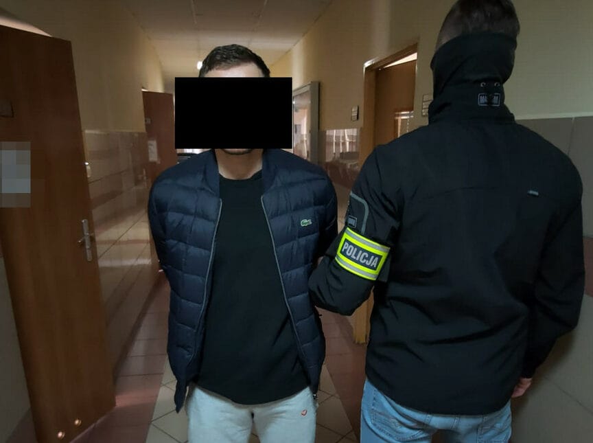 Policjant aresztujący podejrzanego, ma założone ręce i widoczną opaskę POLICJI.