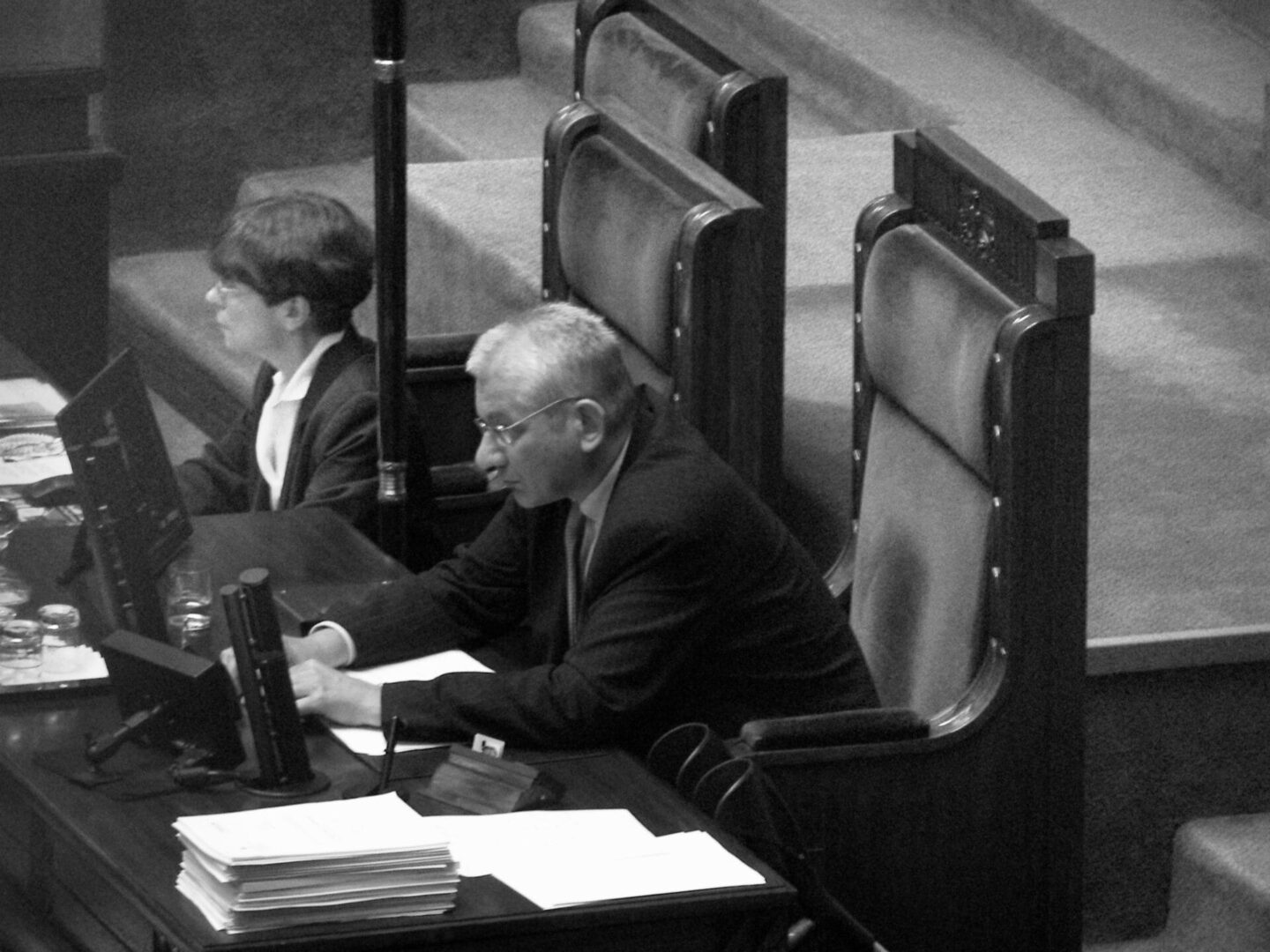 Mężczyzna w garniturze siedzi przy biurku podczas sesji parlamentarnej i przegląda dokumenty.