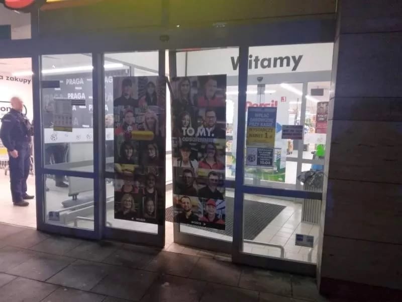 Widok wejścia do sklepu nocą z napisem "Witamy" i plakatami promocyjnymi.