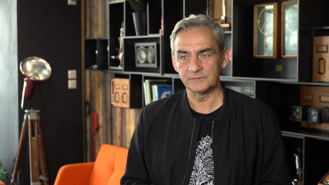 Portret mężczyzny, siwe włosy, poważny wyraz twarzy. Ma na sobie czarną kurtkę.