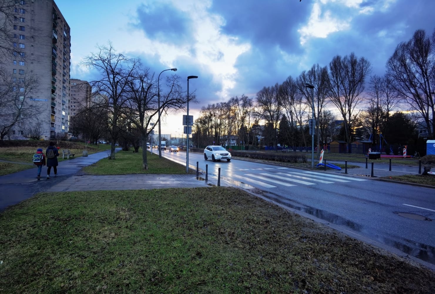 Piesi i samochody na ulicy miasta o zmierzchu, w pobliżu budynków mieszkalnych.