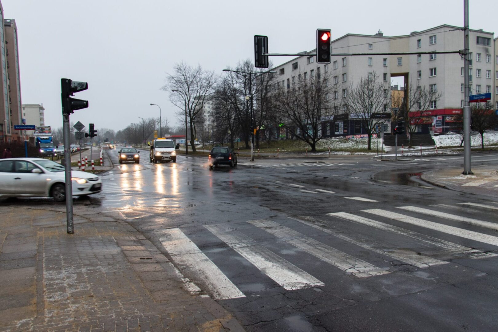 Mokre skrzyżowanie ulic miejskich z przejściem dla pieszych, sygnalizacją świetlną i samochodami.