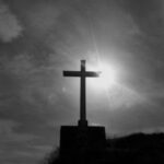 Sylwetka krzyża na szczycie wzgórza, za którym świeci słońce.