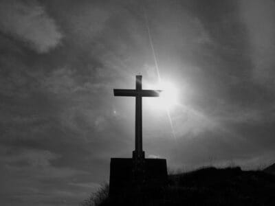 Sylwetka krzyża na szczycie wzgórza, za którym świeci słońce.