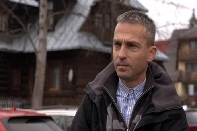 Poważny mężczyzna w ciemnej kurtce, na zewnątrz, niedaleko drewnianego domu.