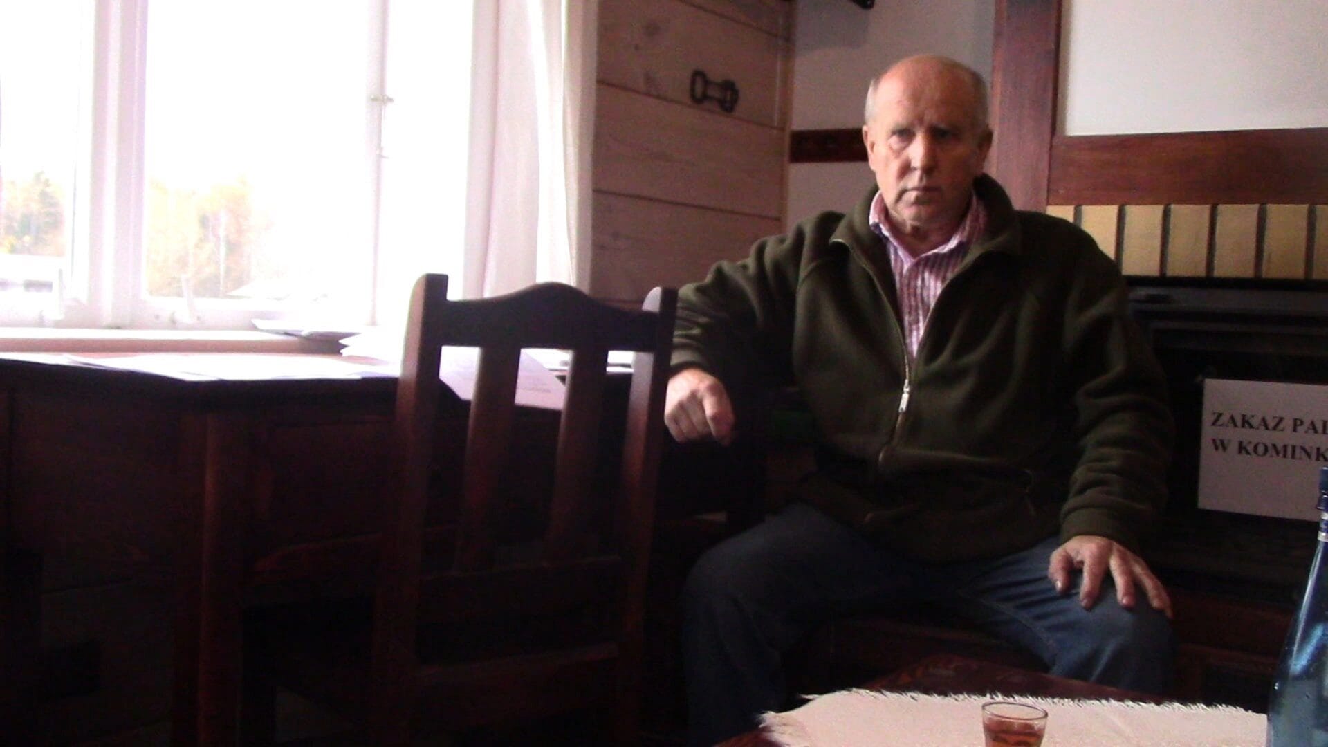 Starszy mężczyzna siedzi na krześle z ciemnego drewna, przed kominkiem. Widoczny znak „Zakaz palenia w kominku”.